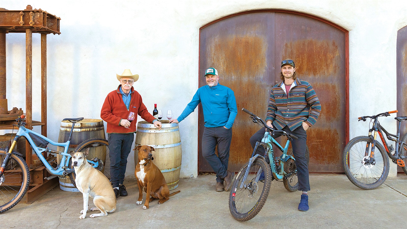 Randy, Mike and Alex Dunn relaxing with glasses of red wine outside with two dogs; Alex is on a bike.